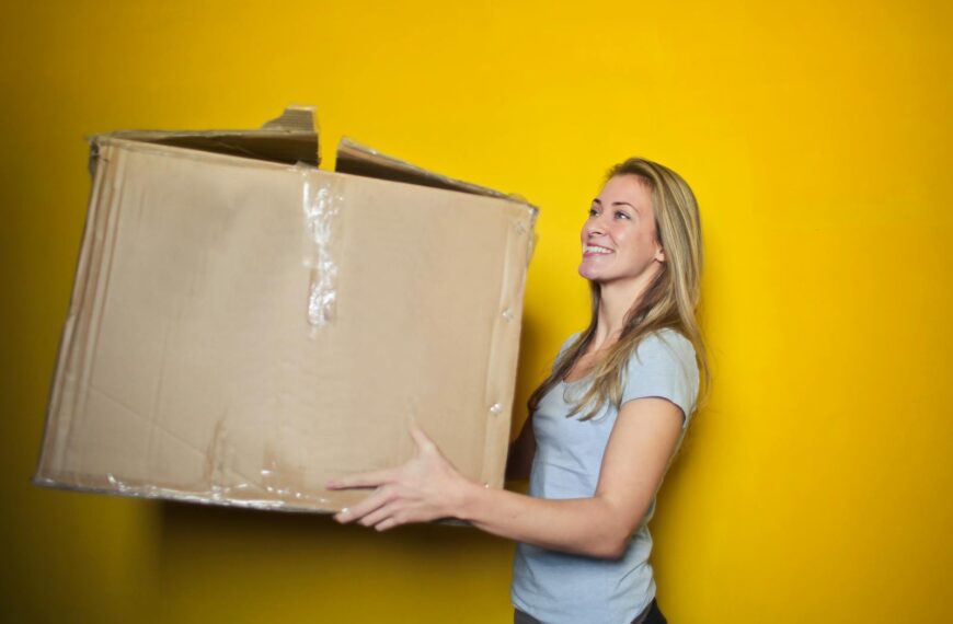 A woman carrying a large cardboard box against a vibrant yellow background, smiling.
