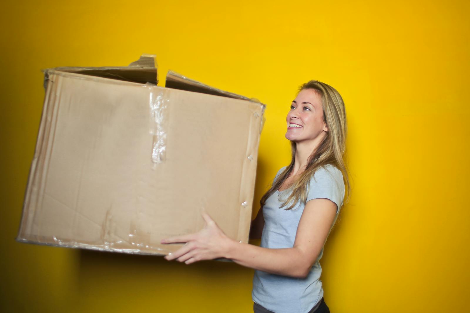 A woman carrying a large cardboard box against a vibrant yellow background, smiling.