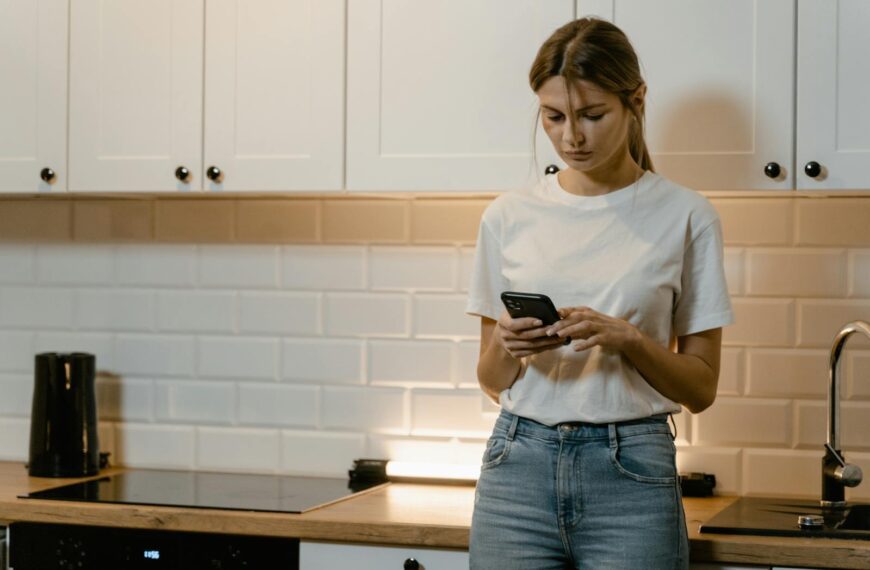 Young woman in kitchen using smartphone, looking focused and thoughtful.