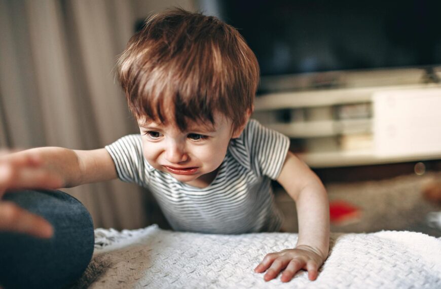 A young child in a striped shirt reaching out with a tearful expression indoors.