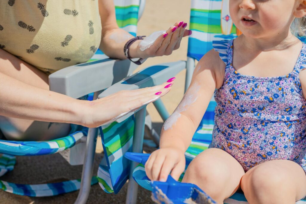 A mother applies sunscreen to her young daughter on a sunny beach day with colorful beach chairs.