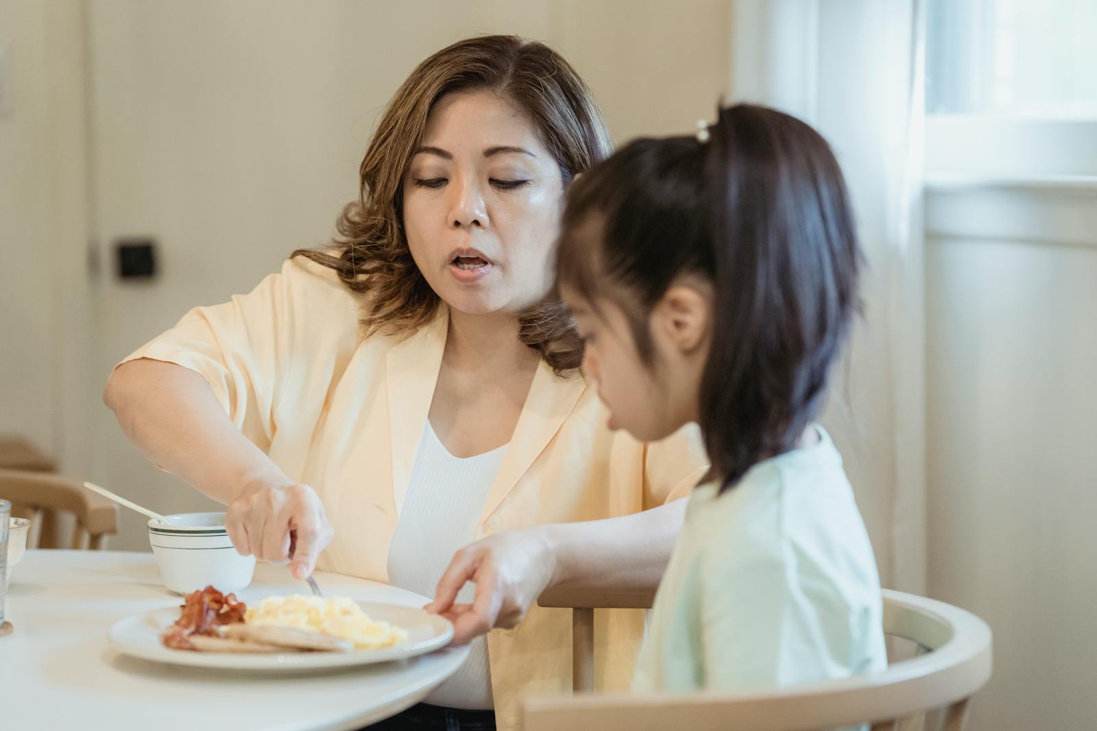 A loving mom feeds her daughter breakfast in a cozy kitchen setting.