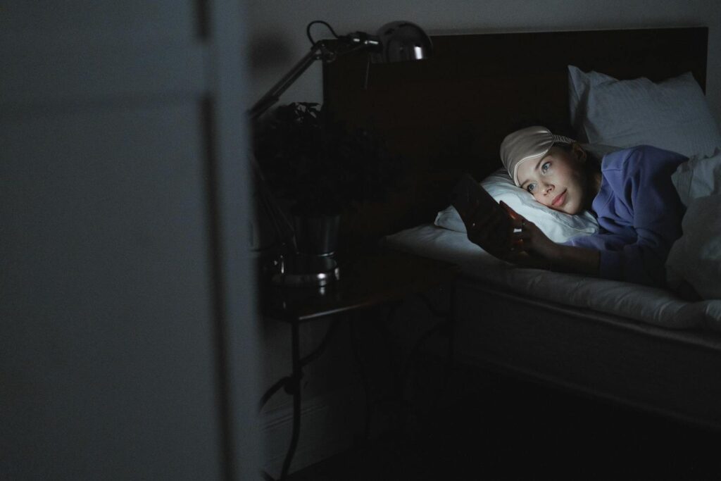 A woman in bed using a smartphone at night, her face illuminated by the screen's light.