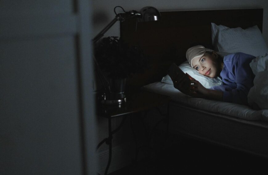 A woman in bed using a smartphone at night, her face illuminated by the screen's light.