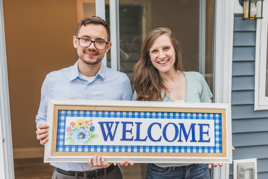 A cheerful couple stands at their new home entrance holding a welcome sign.