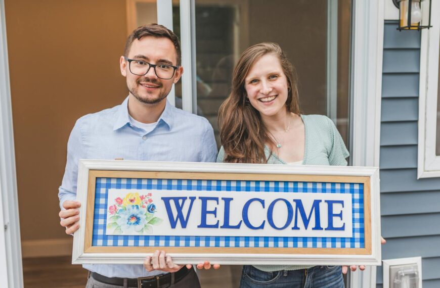 A cheerful couple stands at their new home entrance holding a welcome sign.
