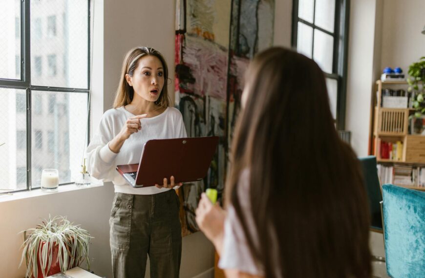 Frustrated mother working on a laptop while talking to her daughter indoors.