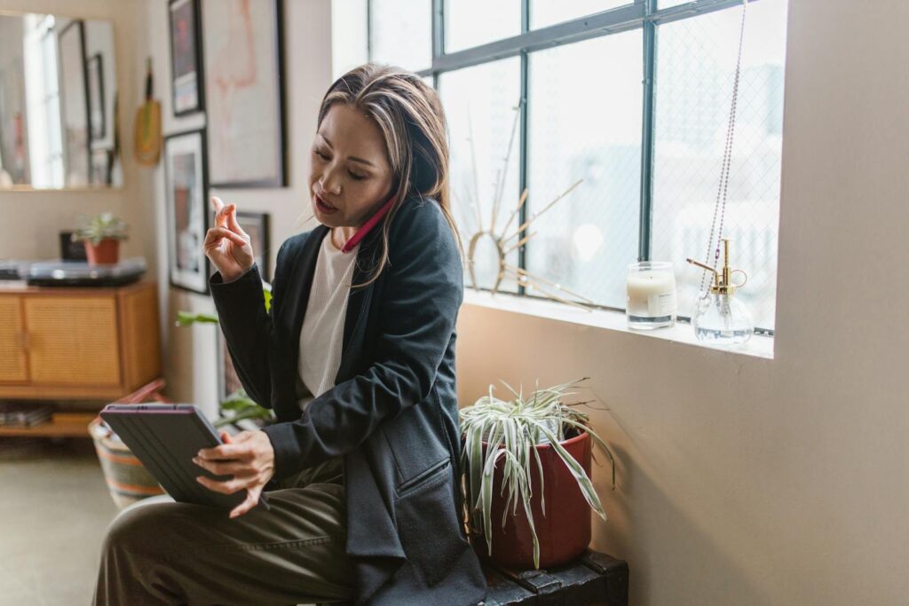 Asian woman multitasks at home using a tablet and phone in a cozy setting.