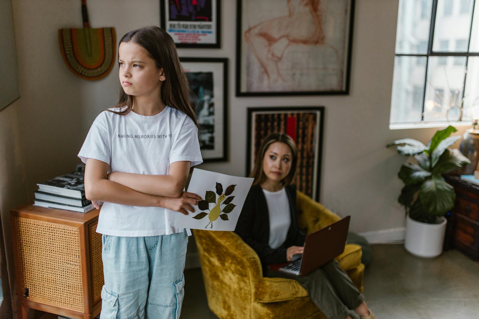 A mother works on a laptop while her daughter holds artwork in a cozy home office.
