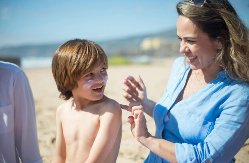 A woman applying sunscreen to her smiling son on a sunny beach day, promoting skin care and safety.