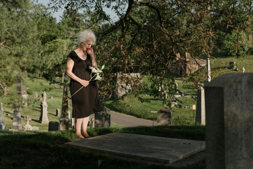 A senior woman standing in a cemetery, holding flowers and expressing grief.
