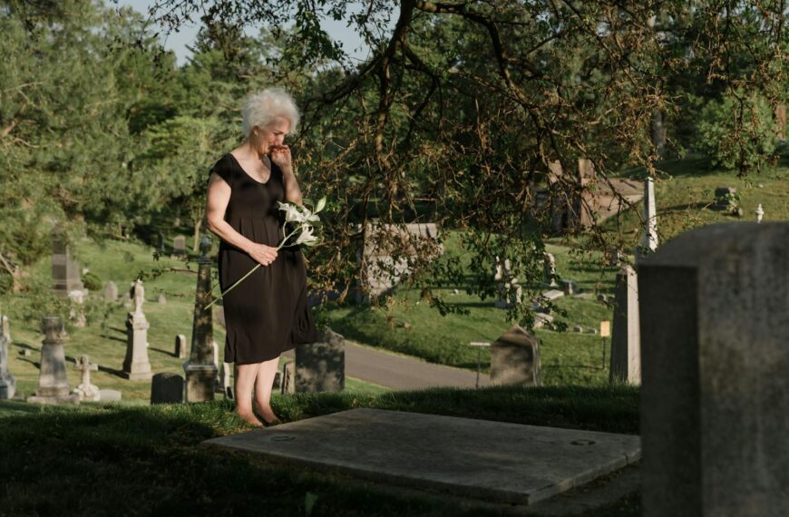 A senior woman standing in a cemetery, holding flowers and expressing grief.