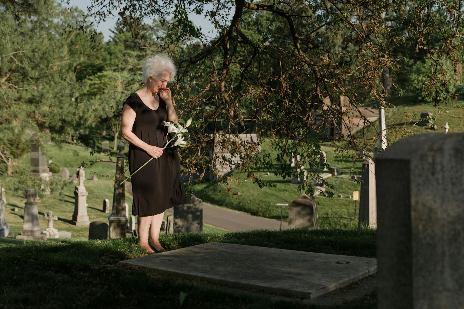 A senior woman standing in a cemetery, holding flowers and expressing grief.