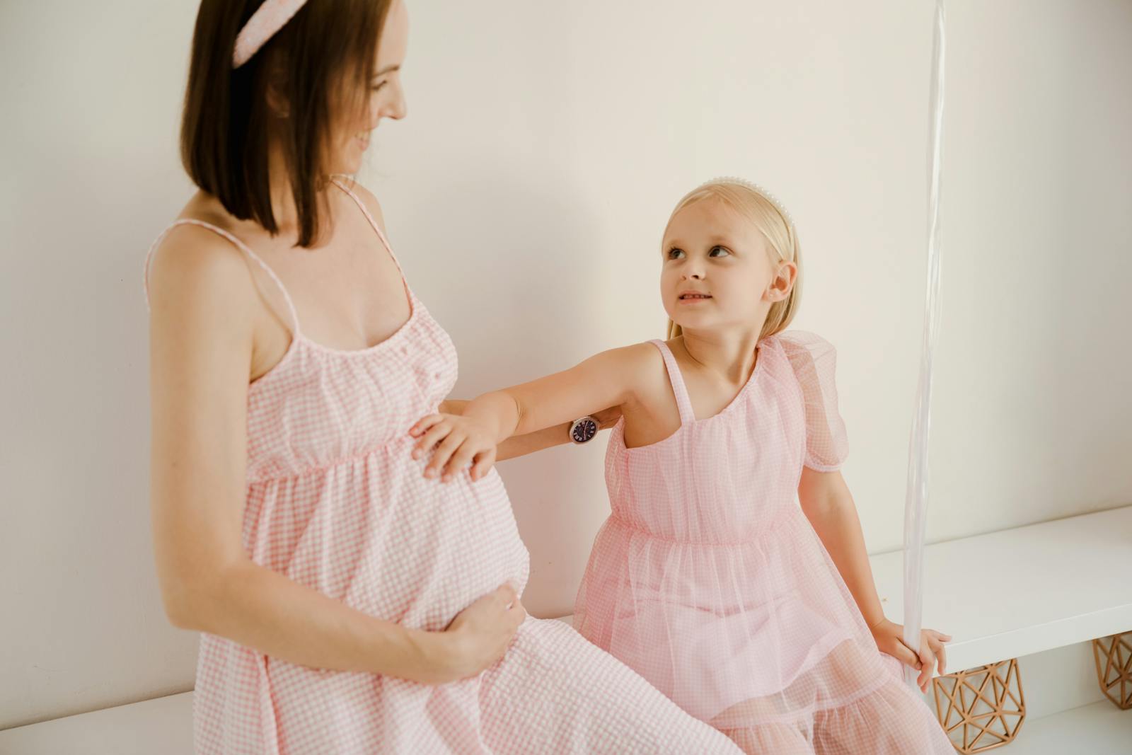 A young girl interacts with her pregnant mother, both wearing matching pink dresses indoors.