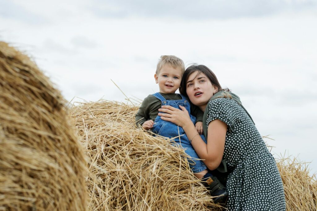 A mother and child enjoying a moment together on a haystack in a serene countryside setting.
