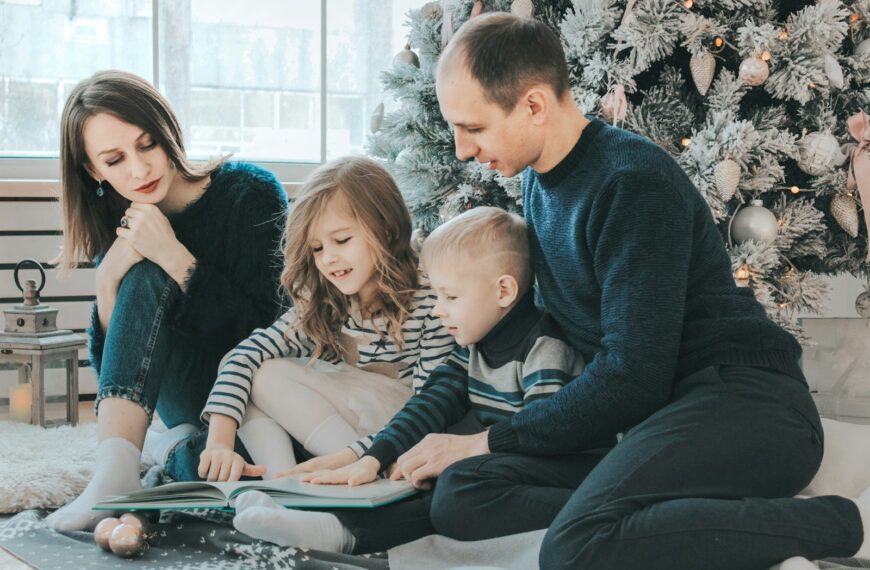 girl and boy reading book sitting between man and woman beside Christmas tree