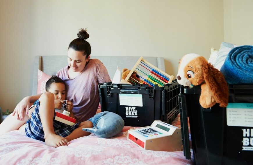 girl in pink shirt sitting on bed beside brown teddy bear