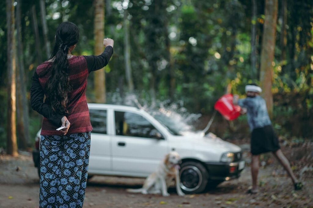 A family scene of washing a car outdoors in a wooded area. Includes a dog.