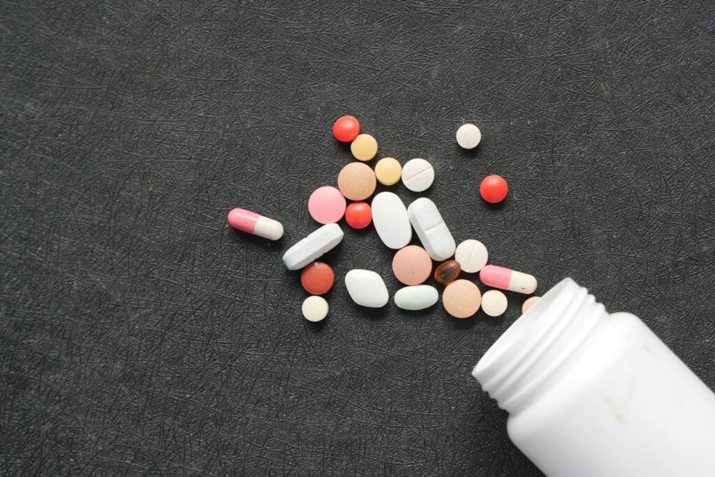 Close-up of diverse pills and capsules spilled from a white bottle on a dark textured surface.