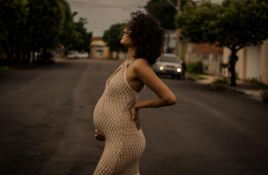 Smiling pregnant woman with curly hair in a beige dress standing outdoors on a street.