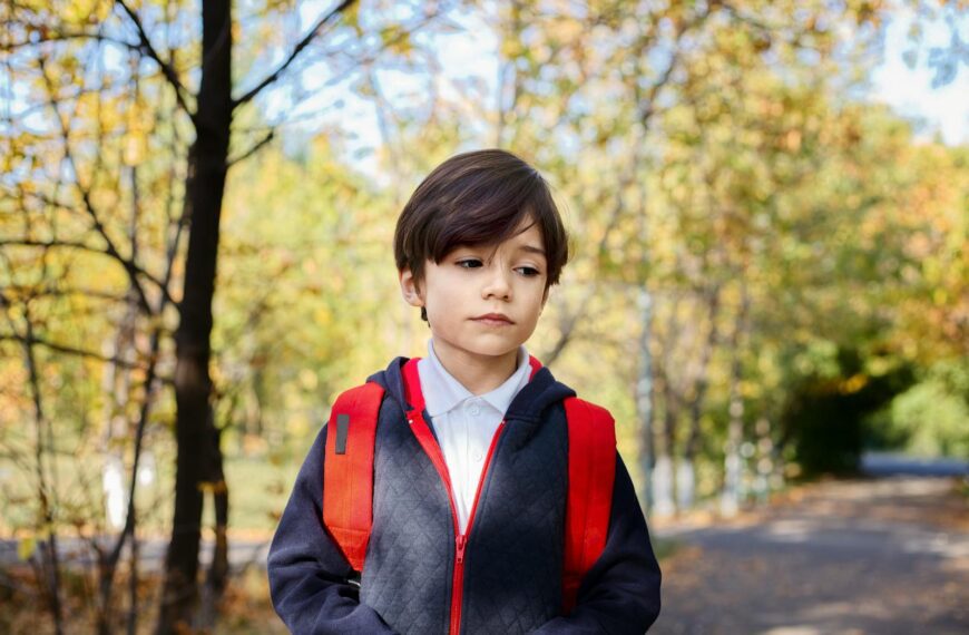 Pensive little boy in school uniform with red backpack looking down while standing in autumn park after studies