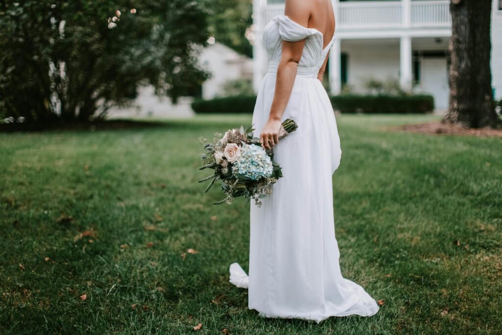 A bride in a white gown stands outdoors holding a bouquet in a lush garden setting.