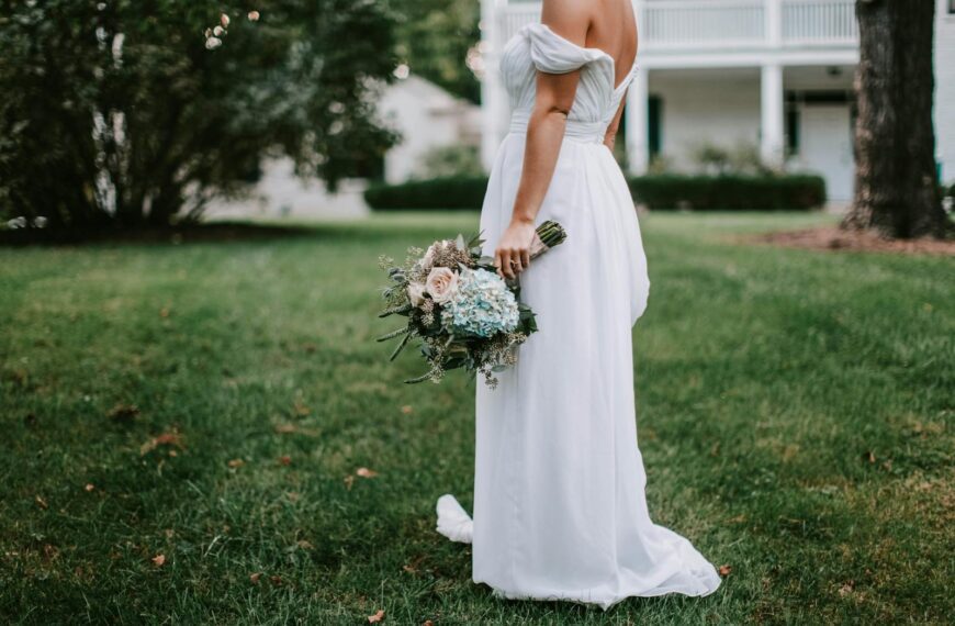 A bride in a white gown stands outdoors holding a bouquet in a lush garden setting.
