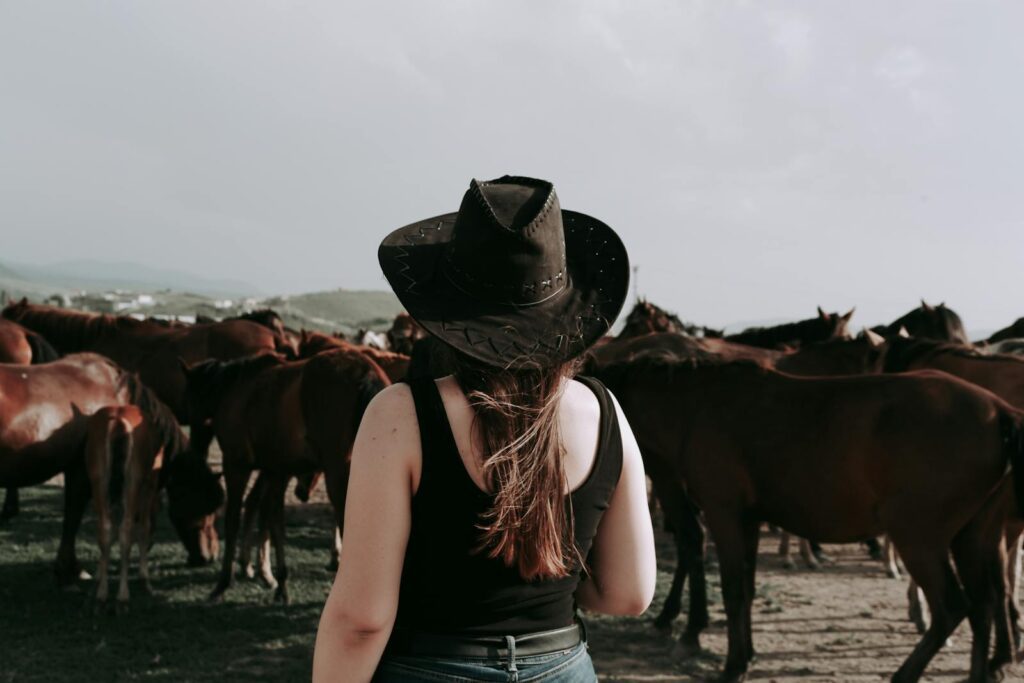A woman in a cowboy hat watching a herd of horses outdoors on a sunny day.