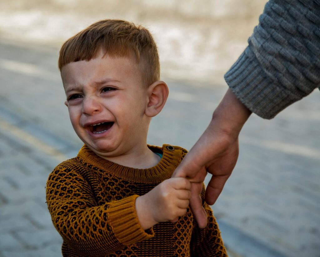 A crying toddler in a sweater holds an adult's hand outdoors, expressing emotion.