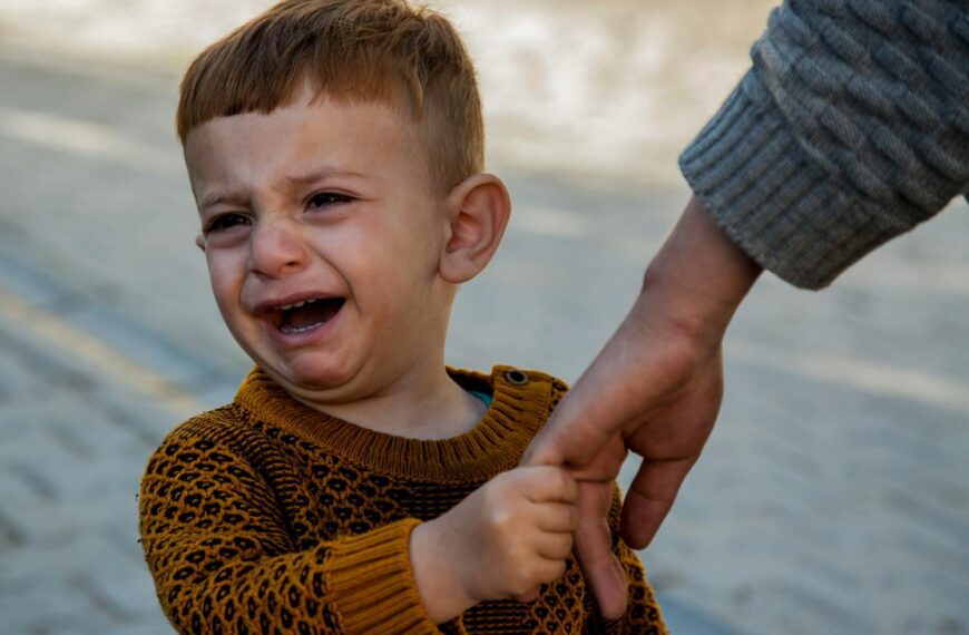A crying toddler in a sweater holds an adult's hand outdoors, expressing emotion.