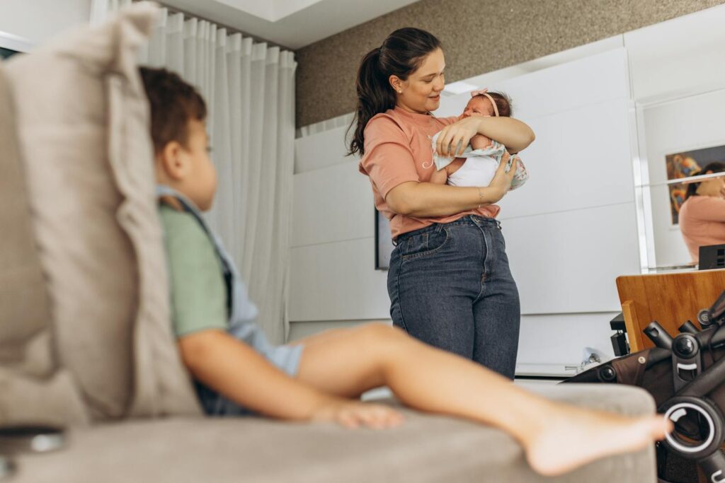 A mother lovingly holds her newborn while a toddler sits on the couch in a cozy living room setting.