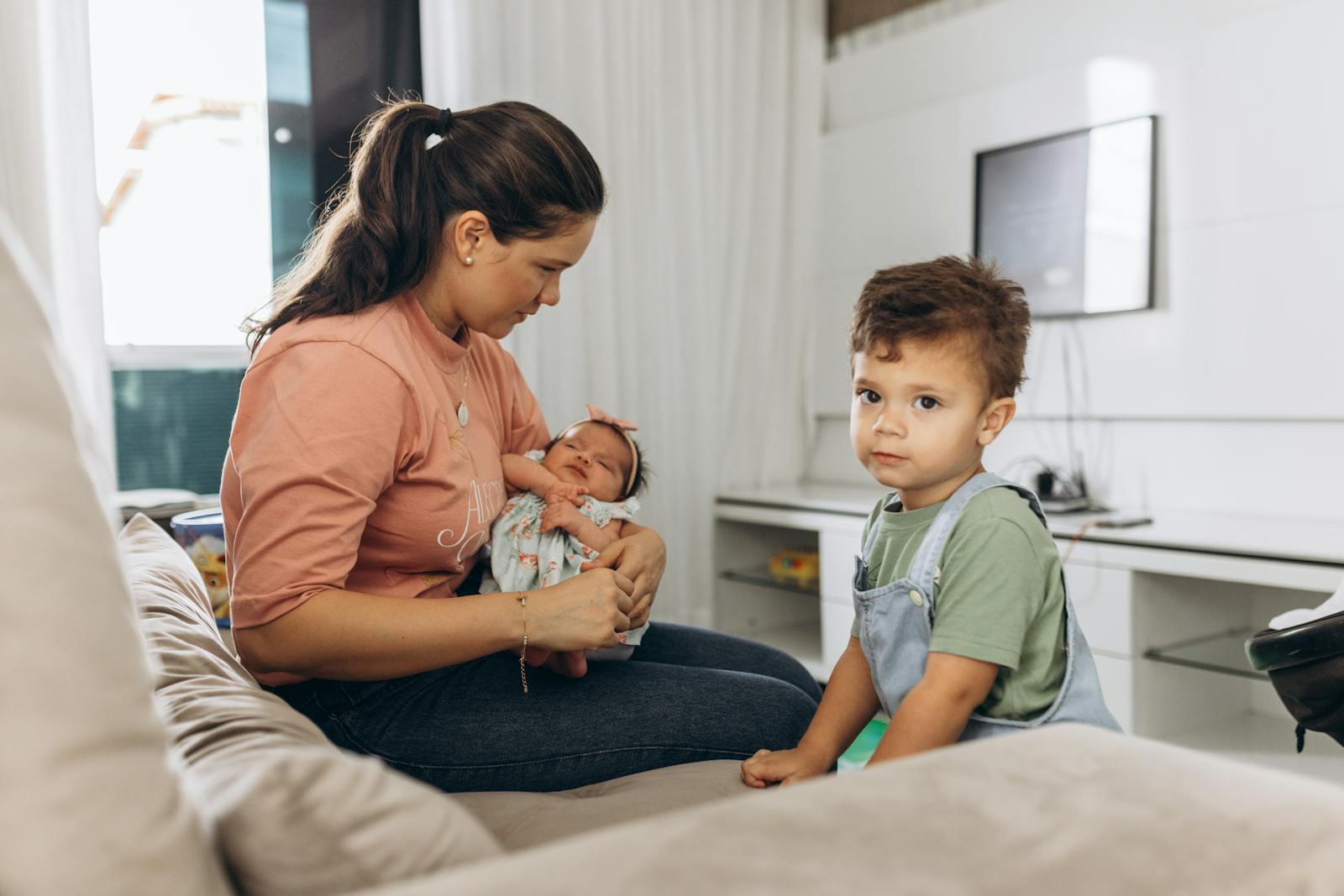 A mother sitting on a sofa with her baby and young son in a bright living room setting.