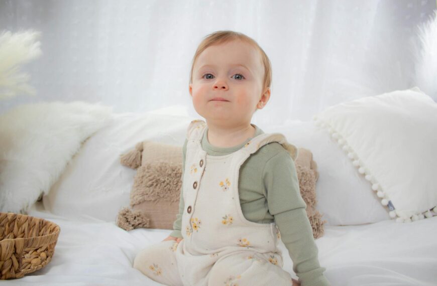 Charming baby in floral overalls sitting on a bed, exuding serenity and cuteness.