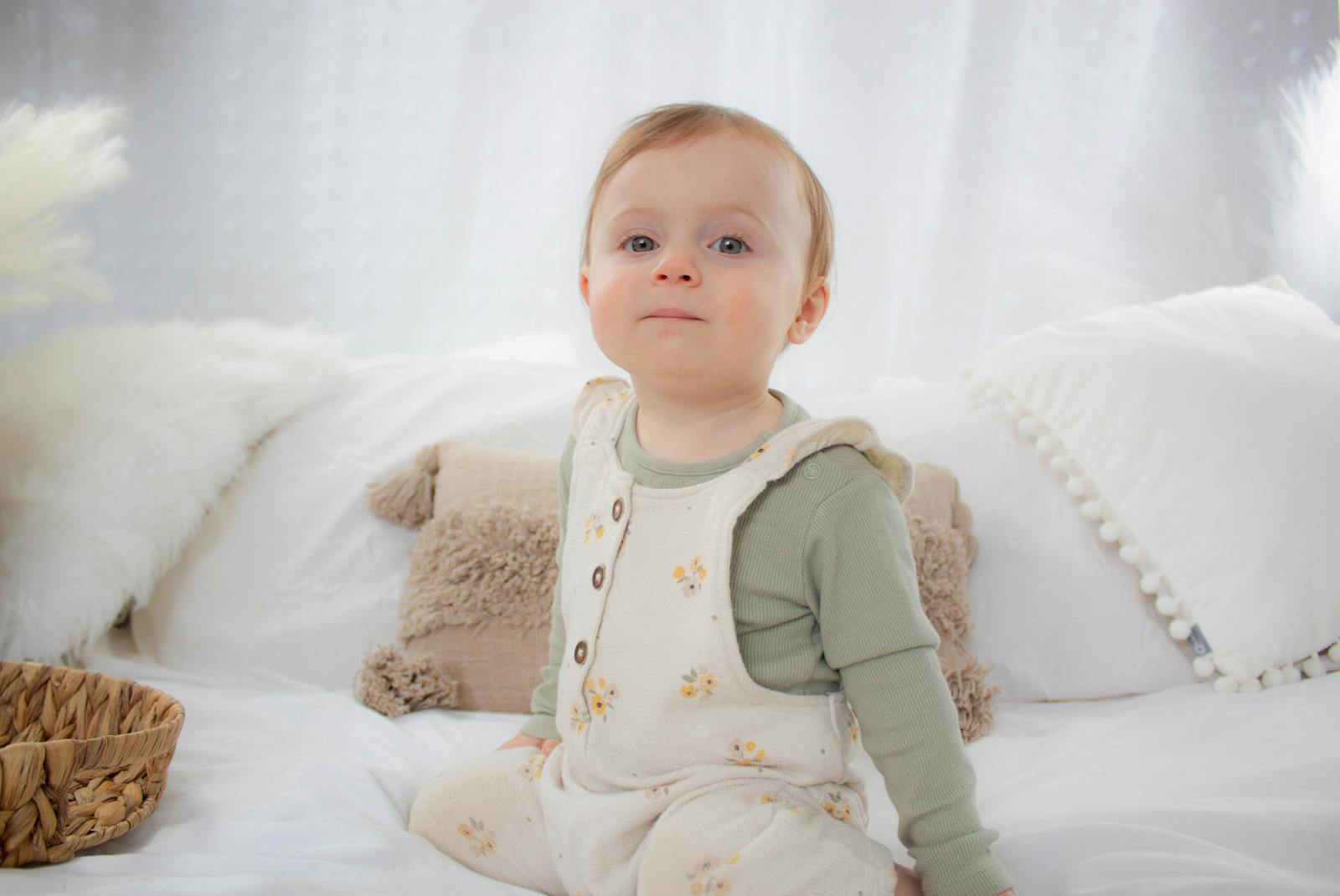 Charming baby in floral overalls sitting on a bed, exuding serenity and cuteness.