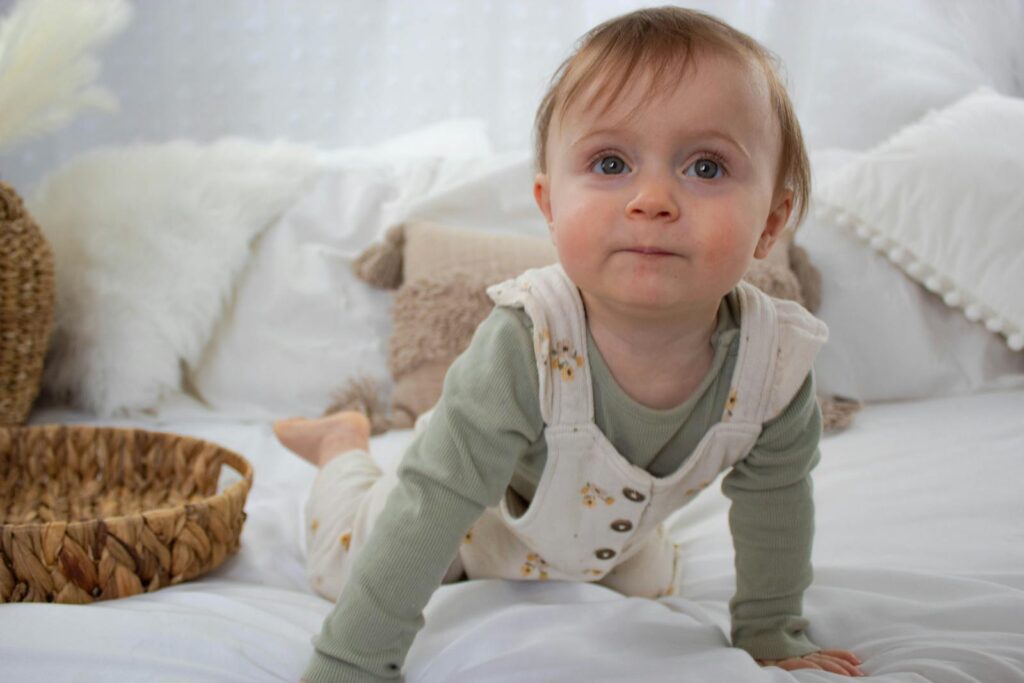 Cute baby in overalls crawling on a soft white bed, with a wicker basket nearby.
