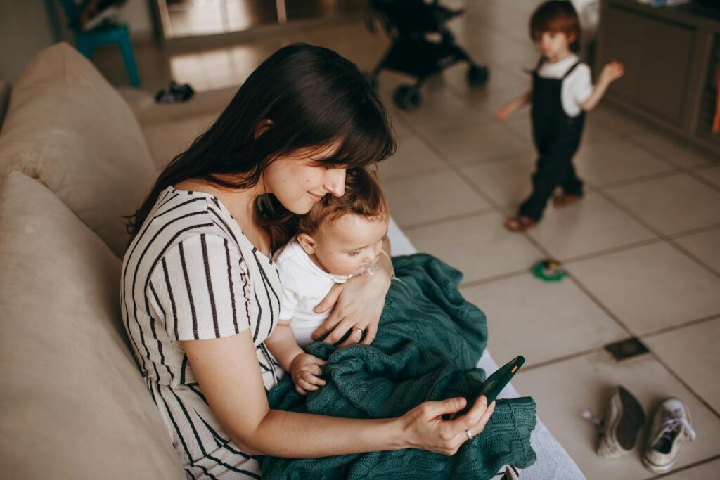 A mother lovingly holds her baby while using a smartphone indoors, with a child in the background.