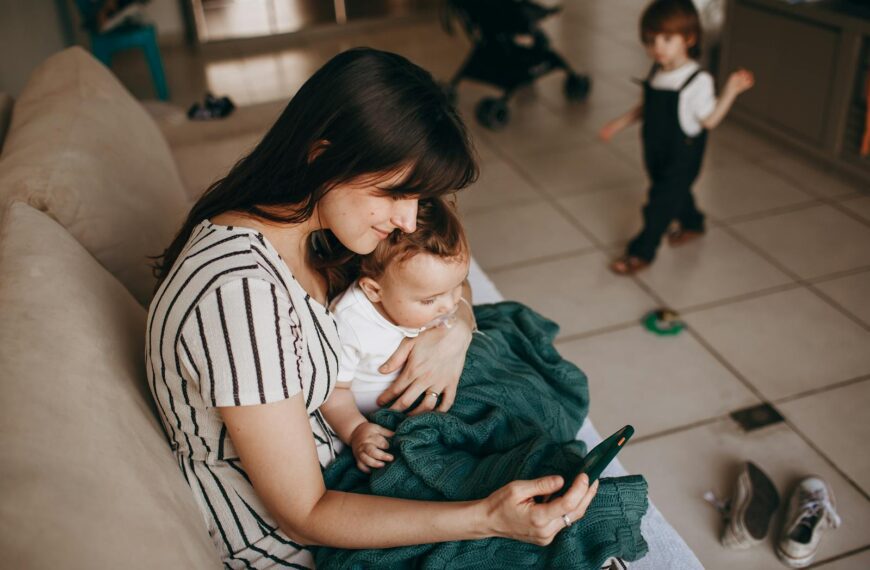 A mother lovingly holds her baby while using a smartphone indoors, with a child in the background.