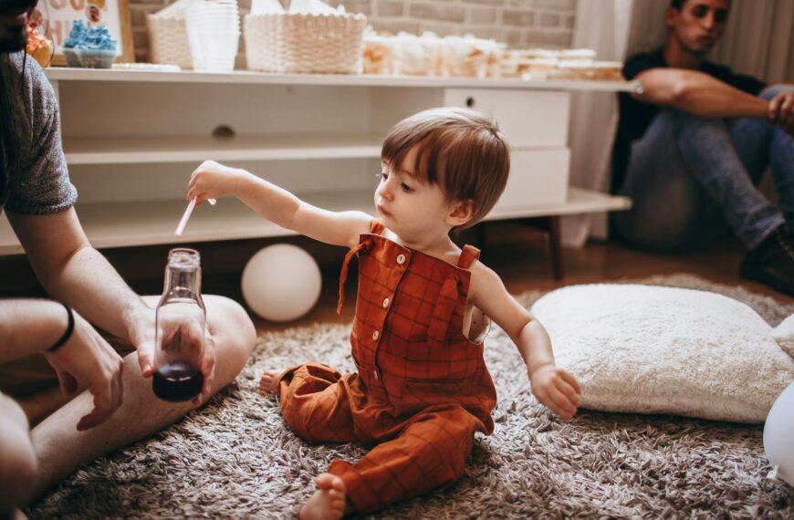 A toddler in an orange romper plays with a bottle indoors. Cozy family setting.