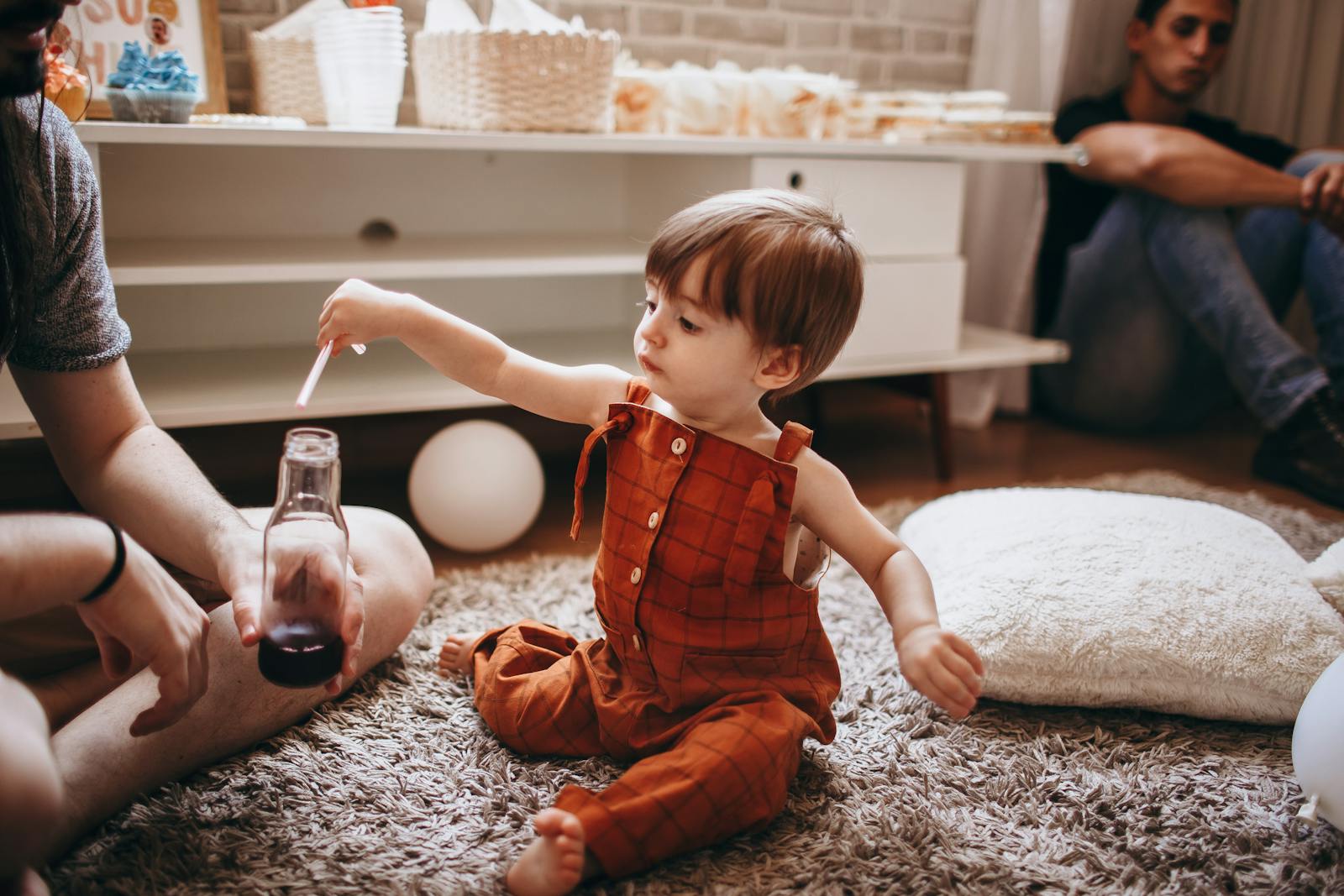 A toddler in an orange romper plays with a bottle indoors. Cozy family setting.