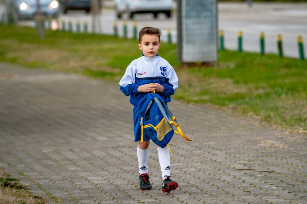 Young boy in sports attire walking on a path outdoors, holding a bag.