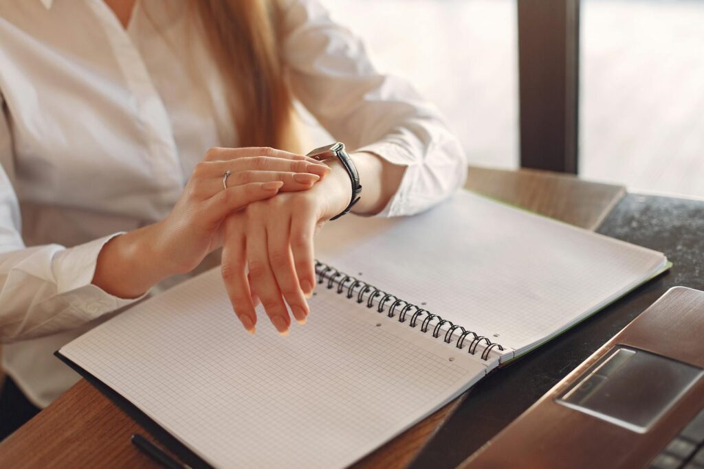 Woman in formal attire reviewing schedule with notebook, checking time on a smartwatch at office desk.