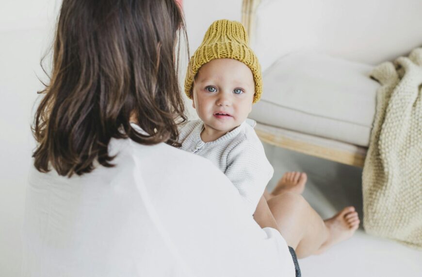 A mother lovingly holds her baby wearing a yellow beanie in a cozy indoor setting.