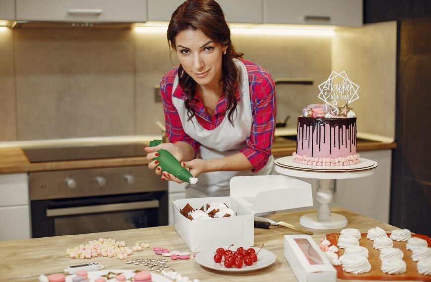 A woman in an apron decorates a cake with icing in a contemporary kitchen setting.