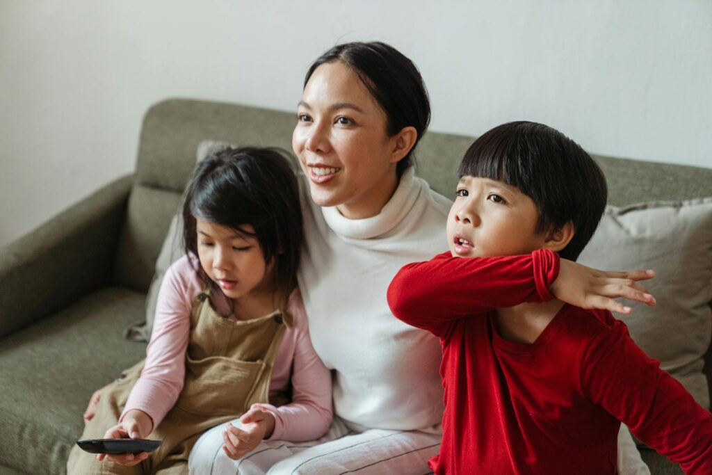 Positive Asian mom and little children watching interesting cartoon on TV while spending day together on comfortable couch