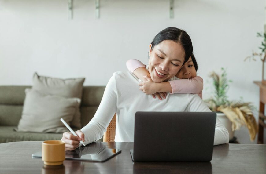 A mother works from home with her daughter embracing her, showing love and family connection.