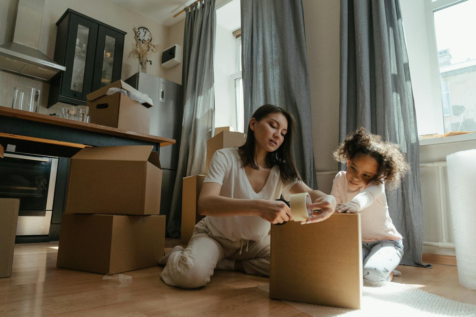 A mother and daughter pack boxes together in a sunny kitchen during a home move.
