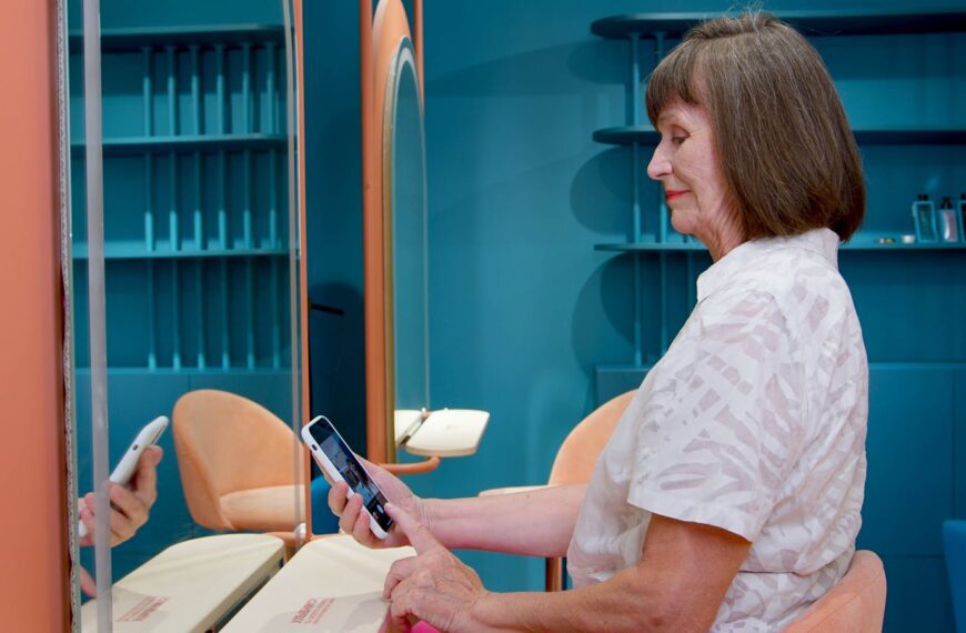 A senior woman sitting in a salon uses her smartphone, reflected in the mirror.