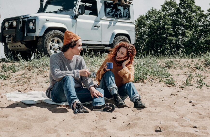 Young couple sitting on a sandy beach, enjoying coffee with a jeep in the background.