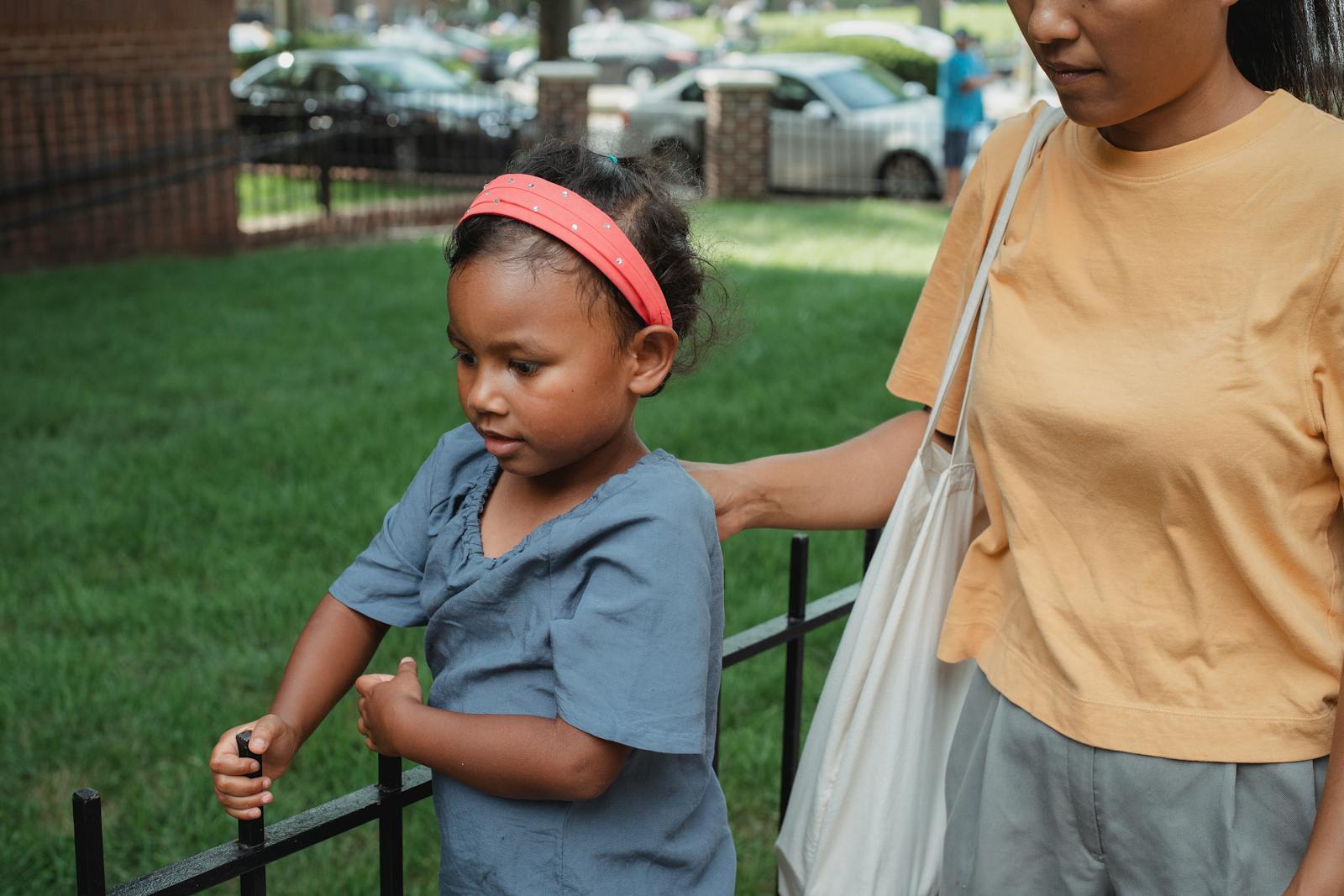Crop anonymous ethnic mother walking with little daughter near metal fence on street in daytime