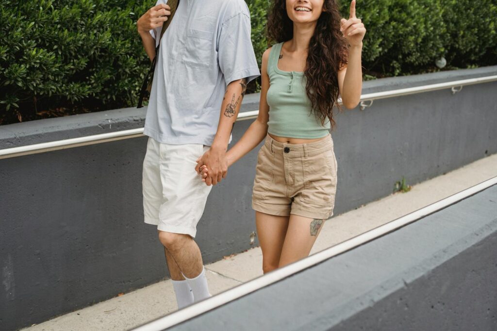 Crop unrecognizable couple holding hands and walking on city street against green plants in daytime